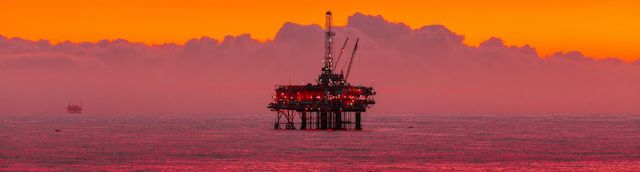 Photo by Arvind Vallabh. Photo depicts an oil rig with red lights in the middle of a purple ocean with ominous red purple clouds and a vivid orange sky. An oil spill glows red in the foreground.